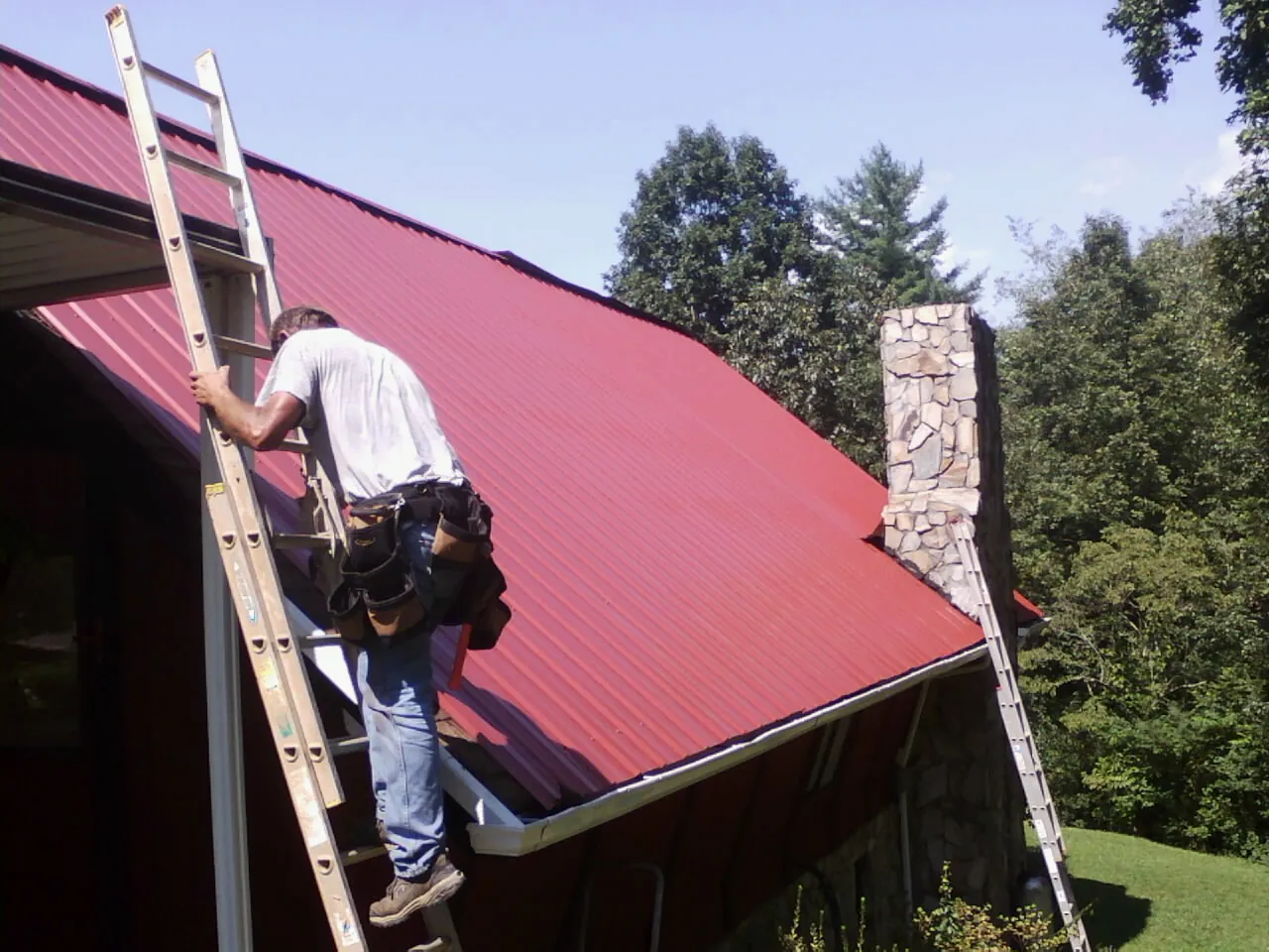 Roofer on a ladder beside a red metal roof and stone chimney — Soffit & Fascia Repair by Roofing Brothers in Benson